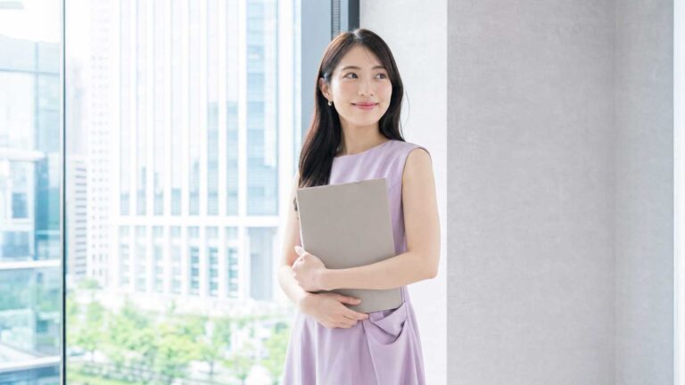 Professional woman in a lavender dress holding a gray binder, standing by large glass windows with a cityscape. (informative)