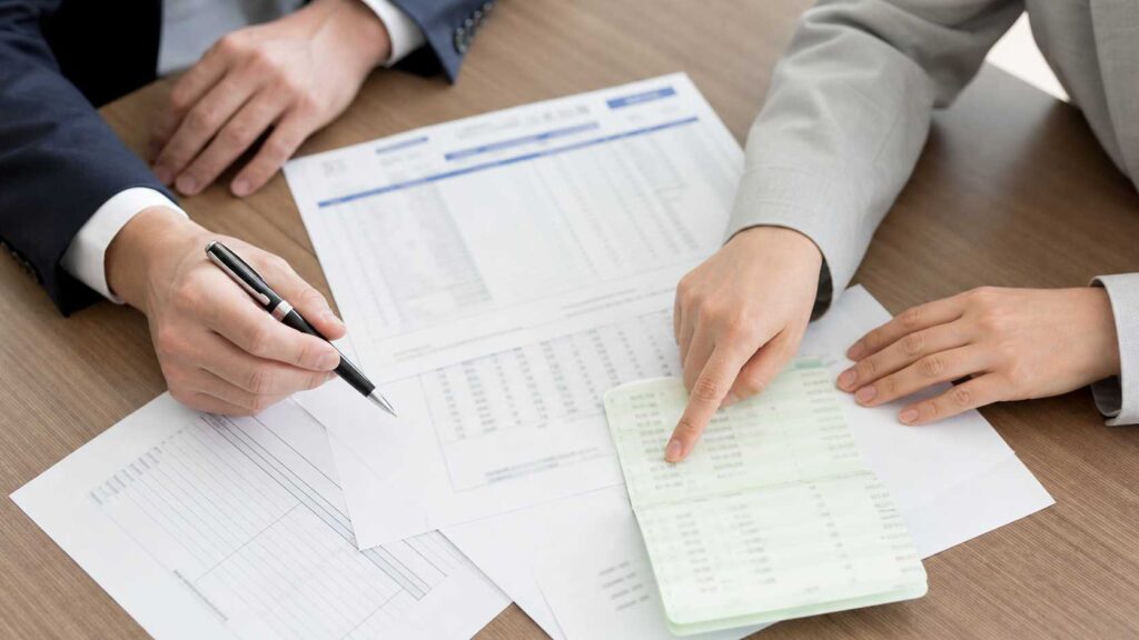 Two professionals review financial documents on a wooden table, one writes with a pen while the other points to a receipt on the sheet area.