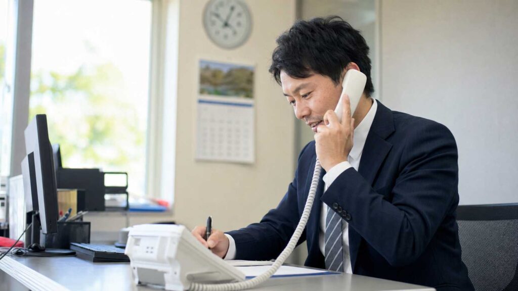 Businessman in a navy suit sits at a desk, talking on a landline phone and taking notes on a paper.