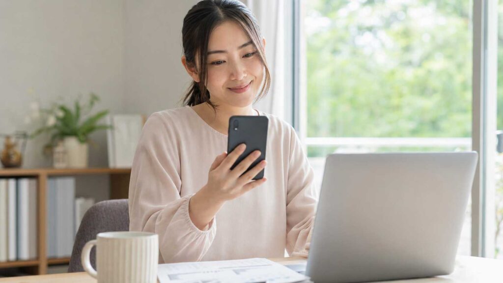 Woman seated at a desk in a bright home office, looking at her smartphone with a subtle smile, laptop open nearby.
