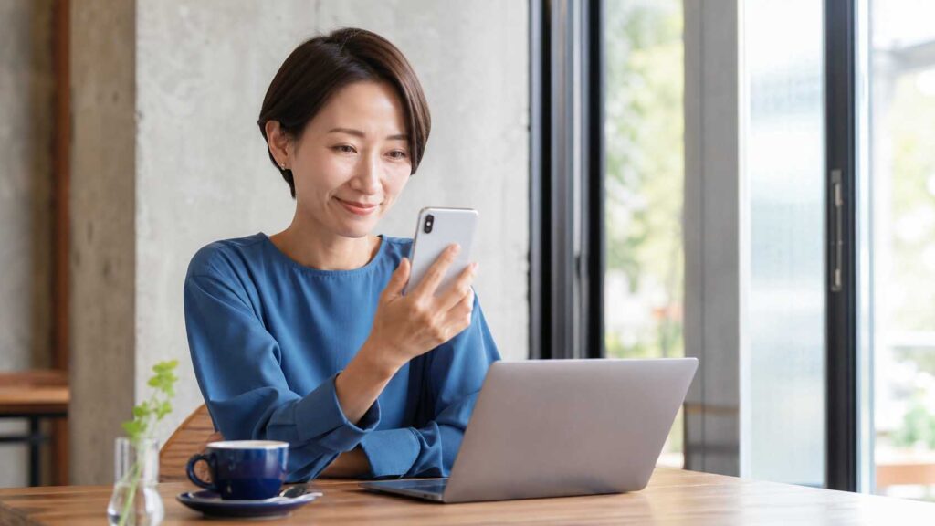 Woman in a blue blouse sits at a wooden table, looking at her smartphone with a laptop nearby in a bright cafe environment.