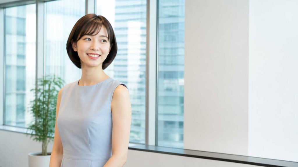 Asian woman in a sleeveless pale blue dress standing by a large office window, smiling softly at the left side of the frame with city buildings in the background.