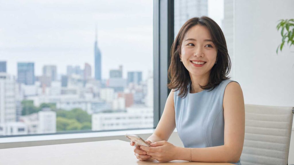 Woman in a light blue sleeveless dress sits at a desk by a window, smiling while holding a smartphone.